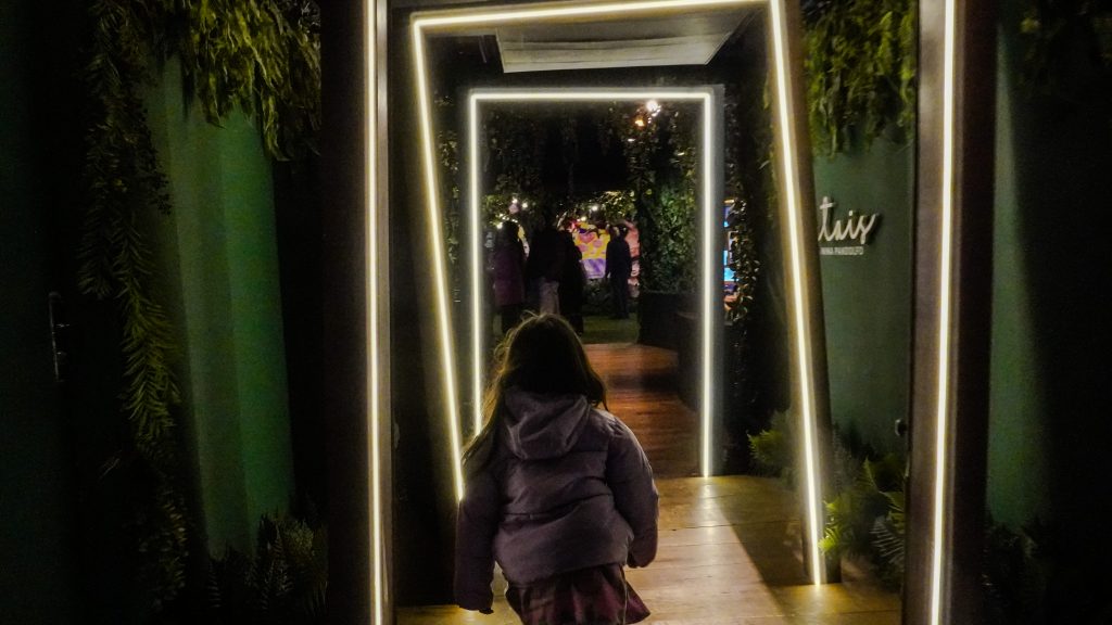 Criança de costas atravessa uma sequência de portais retangulares iluminados, cercados por vegetação, durante uma visita de férias à exposição Portais, de Nina Pandolfo.
