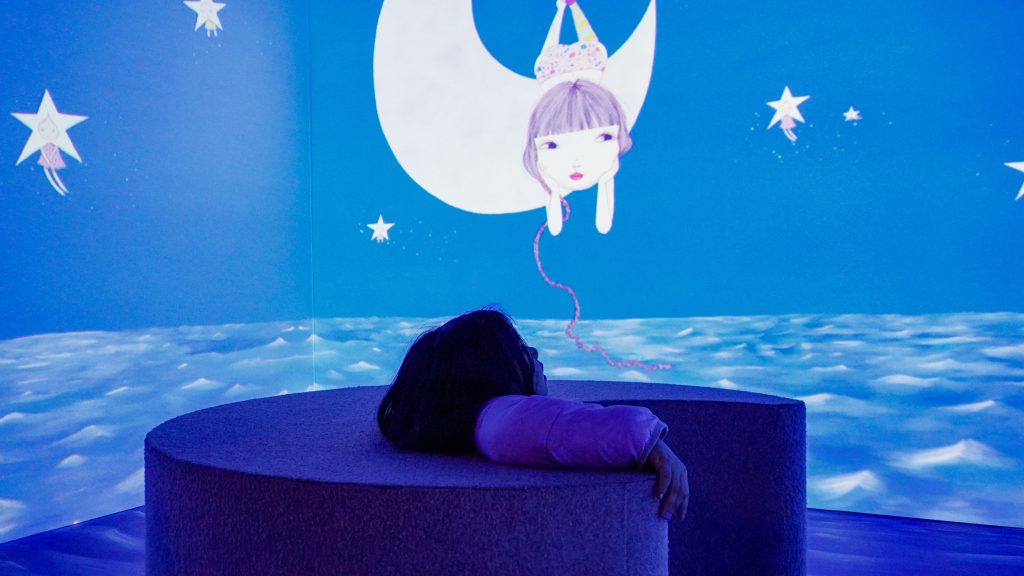 Criança deitada sobre um banco circular durante uma visita de férias, em uma sala escura iluminada por projeções azuis de lua, estrelas, nuvens e uma personagem feminina.