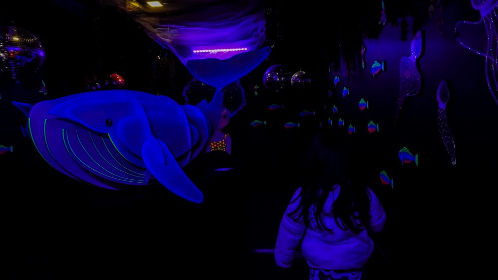 Criança vista de costas durante uma visita de férias, em uma sala escura iluminada por luz ultravioleta, diante de uma grande baleia azul fluorescente, pequenos peixes coloridos e outras figuras marinhas nas paredes.