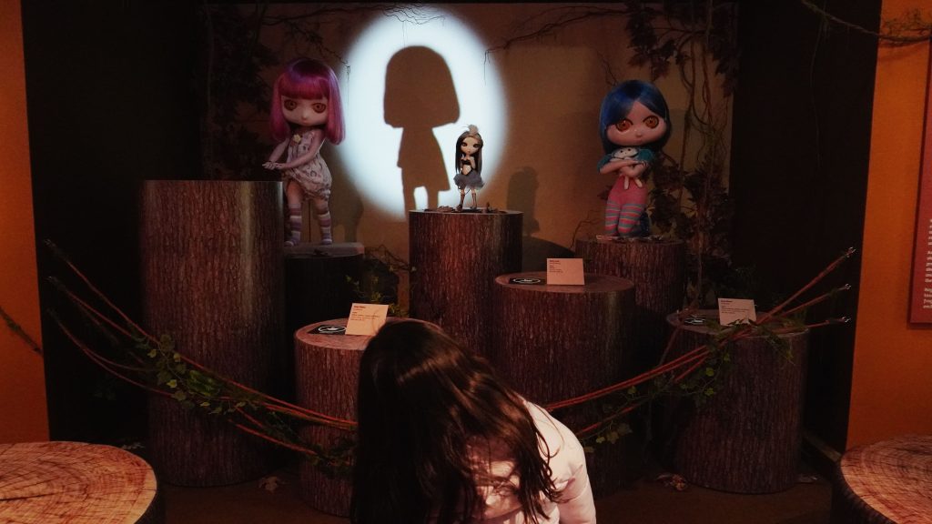 Durante uma visita de férias, uma criança observa três bonecas expostas sobre bases que imitam troncos, em uma sala escura. Um foco de luz projeta a sombra de uma das figuras na parede.