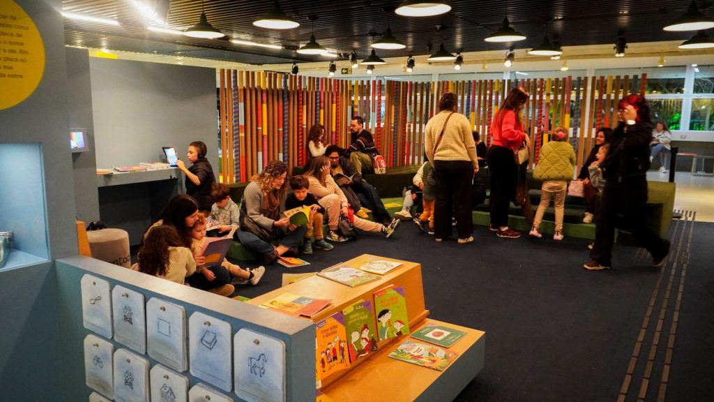 Durante as férias, adultos e crianças sentados em almofadas e bancos leem livros em uma área da exposição. Exemplares de Ruth Rocha estão dispostos em mesas e prateleiras no primeiro plano.