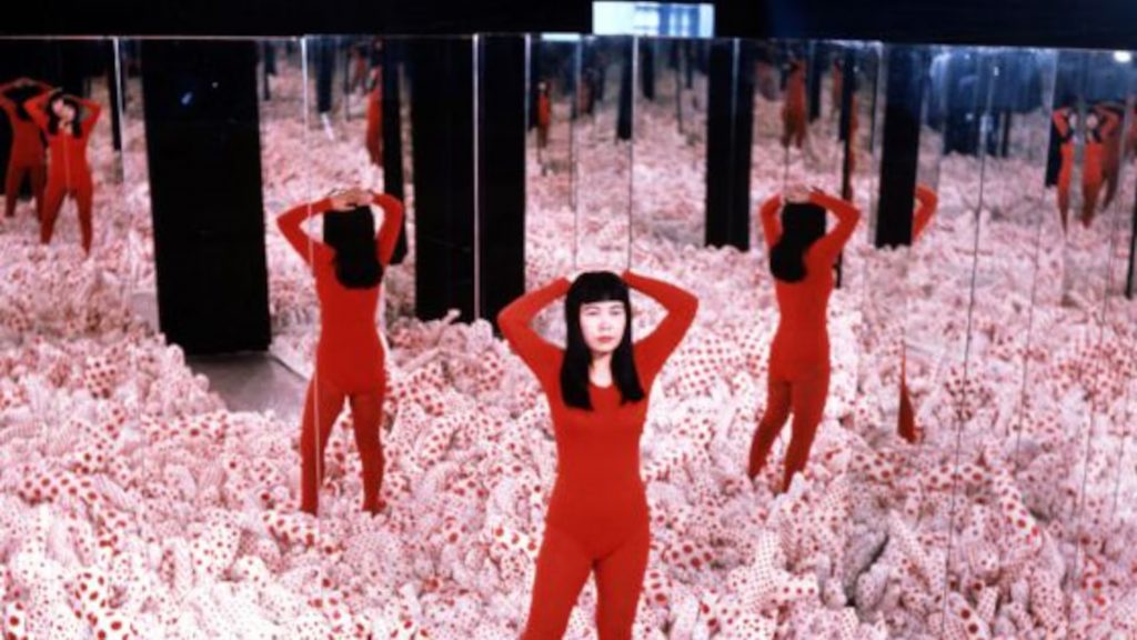 Yayoi Kusama ao lado de um cavalo durante performance ao ar livre, com corpos cobertos por bolinhas