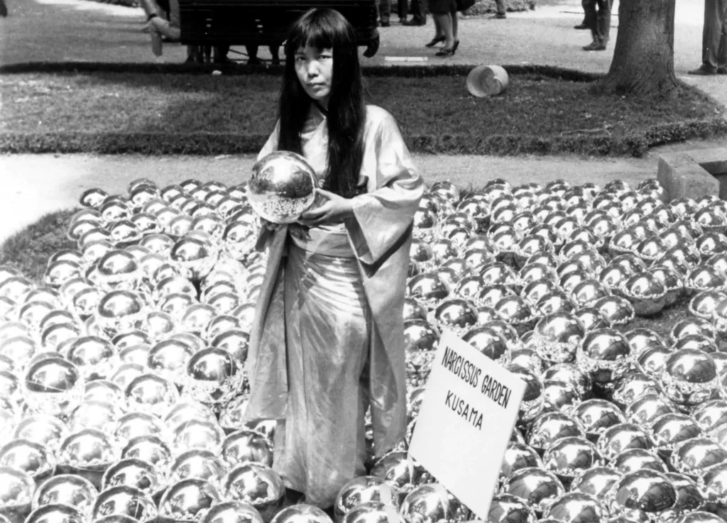 Yayoi Kusama em sala espelhada repleta de formas brancas com bolinhas vermelhas, multiplicadas pelos reflexos.
